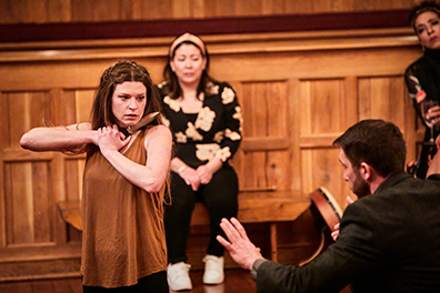Juliet in brown tank top holds a dagger to her throat as Friar Laurence holds up his hands trying to calm her.