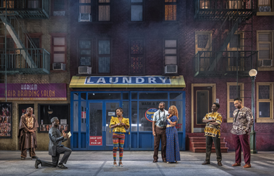 STC-Merry_Wives_Street-091 Characters in various West African-styled clothing (except the Pages, he in common American short-sleeve shirt and pants with binoculars hanging around his neck from birdwatching, and she in floor length blue dress with small yellow X's) watch Ford's contritio in front of a street scene of the salon and laundryomat with tenements on either sice and a street lamp to one side.